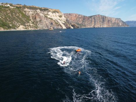 Happy people swim on air mattress behind a high speed water bike. Tourists ride on inflatable watercraft boat. Sea attraction. Water boat skating people on large Inflatable raft. Aerial copter view