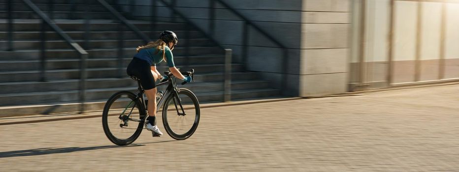 Professional female cyclist in cycling garment and protective gear riding bicycle in city, rushing and passing buildings while training outdoors on a daytime. Urban lifestyle, sports concept