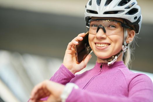Adorable young female cyclist wearing protective helmet and glasses smiling at camera while talking on the phone, standing outdoors on a daytime. Safety, sports, technology concept