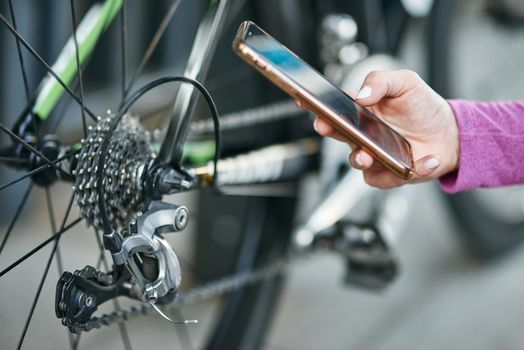 Close up shot of hand of female cyclist holding smartphone while checking her bicycle mechanisms, sprocket and chain on a mountain bike outdoors on a daytime. Safety, sports, technology concept