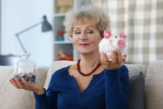 Portrait of senior woman on pension weighing containers with saved money for future. Piggy bank and glass container full of coins. Budget, money concept