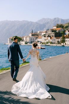 Bride and groom hold hands and walk along the asphalt embankment of Perast. High quality photo