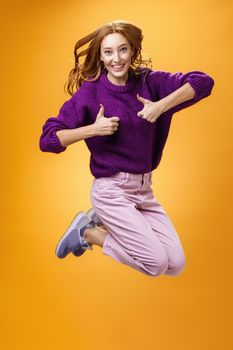 Optimistic and cheerful woman liking promotion lifting thumbs up and jumping from excitement and delighted, having fun recommending good idea smiling thrilled and enthusiastic over orange background. Copy space