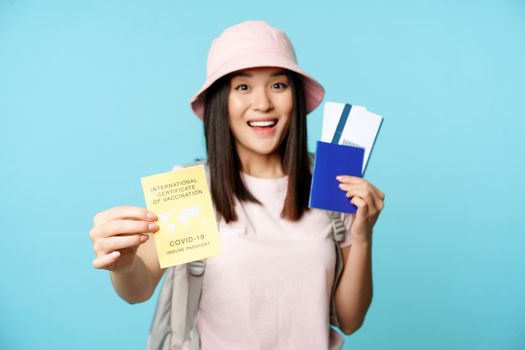 Enthusiastic asian tourist, girl shows covid-19 international vaccination certificate, passport with two tickets, travelling after coronavirus vaccine, blue background.
