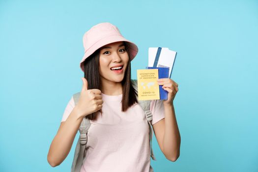 Smiling happy asian tourist, girl shows thumbs up and international vaccination certificate, vaccine passport and tickets, standing over blue background.