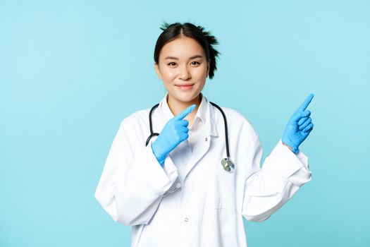 Smiling asian healthcare worker, wearing sterile gloves and uniform, pointing right, demonstrating chart, hospital advertisement, blue background.