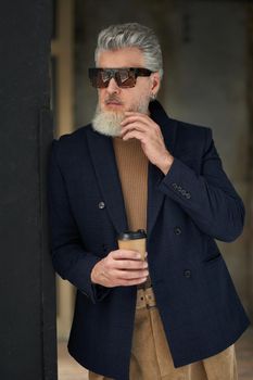 Stylish middle aged man in business casual wear and sunglasses leaning on the wall, holding disposable coffee cup while posing in loft interior. Lifestyle, people concept