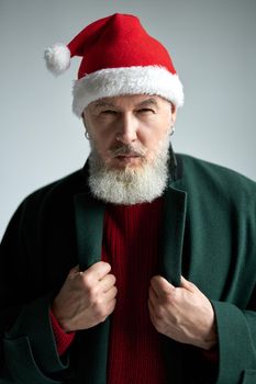 Focused middle aged man with Christmas hat wearing stylish outfit looking at camera, adjusting his coat while posing isolated over light gray background. Holidays concept