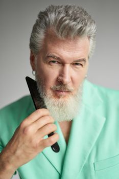 Portrait of attractive gray haired mature man looking at camera, brushing his beard with comb, posing over white background. People, lifestyle concept