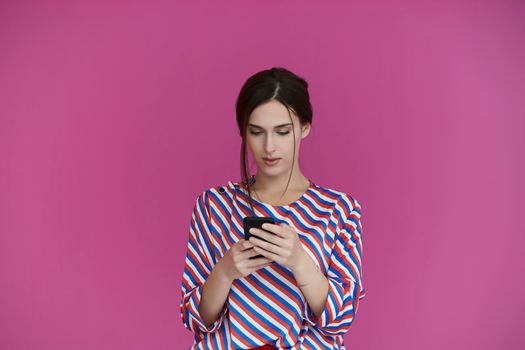 Portrait of young girl with serious face while using smartphone isolated on pink background. Female model chatting or browsing on social networks. Fashion and technology concept