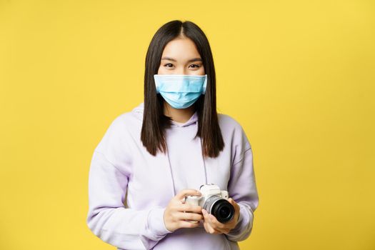 Image of young asian woman in medical face mask, taking pictures on camera, photographer working during pandemic, yellow background.