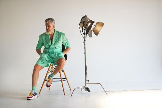 Studio shot of trendy gray haired middle aged man wearing stylish outfit with colorful sneakers looking aside, sitting next to studio spotlight, posing over white background. Fashion, style concept