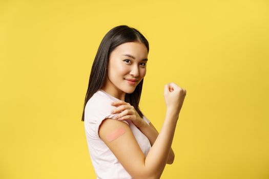 Covid-19 vaccination concept. Smiling asian girl showing patch from coronavirus vaccine on arm, took shot from flu, yellow background.