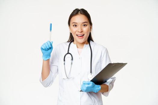 Smiling asian female doctor raising pen up, eureka gesture, holding clipboard, standing in medical uniform over white background.