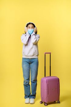 Full length shot of happy smiling asian tourist, girl in headphones, listening music in airport, wearing medical face mask, standing with suitcase, yellow background.