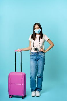 Happy asian female tourist, standing in medical face mask with suitcase and camera, going on tour, vacation abroad during covid-19 pandemic, blue background.