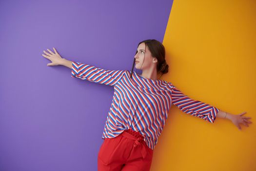 Young female model with open wide arms posing over purple and yellow background. Girl in modern fashionable clothes leaning against wall in the studio with stretched hands. Freedom