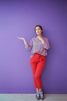 Portrait of happy smiling young beautiful woman in a presenting gesture with open palm isolated on purple background. Female model in modern fashionable clothes posing in the studio