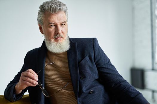 Portrait of stylish serious middle aged man with beard looking at camera, holding glasses while sitting on sofa indoors. Lifestyle, people concept