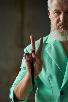 Close up shot of stylish middle aged man with beard wearing colorful outfit looking at camera, showing peace sign while holding sharp barber scissors, posing indoors. Professional occupation concept