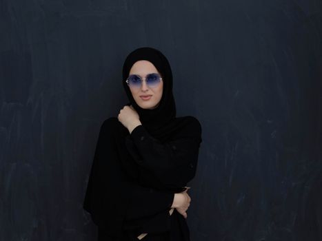 Young muslim in traditional clothes or abaya and sunglasses posing in front of black chalkboard. Arab woman representing modern arabic lifestyle, islamic  fashion and Ramadan kareem concept