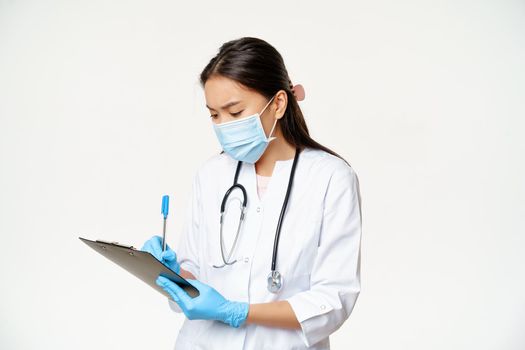 Asian female doctor writing on clipboard, holding pen and paper, standing in face medical mask and rubber gloves, white background.