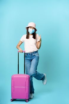 Happy korean woman in medical face mask, thumbs up, posing with cute suitcare, tourist going on vacation, travelling during covid-19 pandemic concept, blue background.