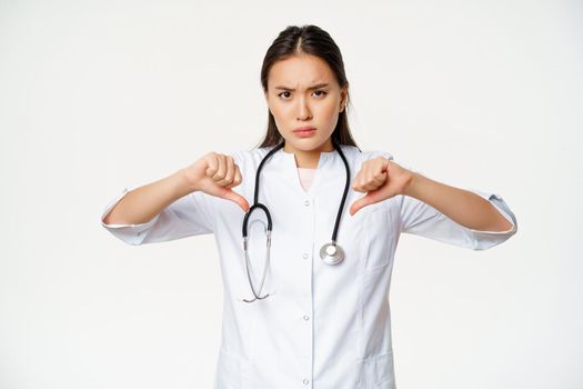Disappointed serious-looking asian female doctor, hospital physician shows thumbs down with disapproval, dislike smth bad, standing over white background.