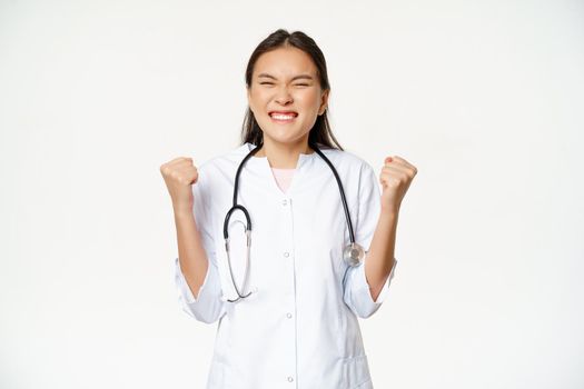 Happy asian intern celebrating. Physician doctor triumphing and rejoicing, say yes with clenched fists pump, standing ecstatic over white background.