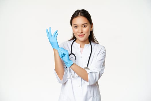 Smiling confident asian female doctor put on medical gloves, getting ready for patient examination, standing in uniform over white background.