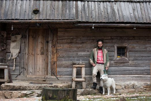 handsome young hipster man standing together with white husky dog in front of old vintage retro wooden house