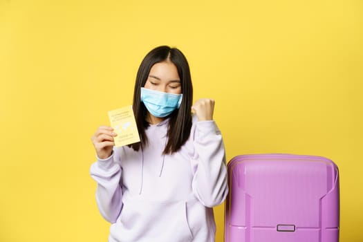 Happy asian girl tourist got covid international vaccination certificate, going on vacation and rejoicing, celebrating her trip, standing in medical mask, yellow background.