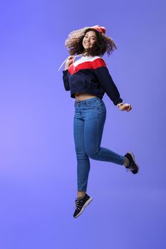 Dreamy and femenine cute girl with curly hair in winter beanie, jeans and sweatshirt jumping over blue background with satisfied carefree smile looking at camera feeling light and joyful.