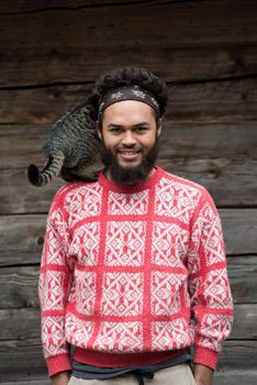 portrait of young hipster man with beard and cat in front of wooden house