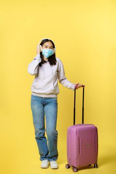 Full length shot happy asian young girl tourist, international traveller in headphone, standing near suitcase and smiling, wearing medical face mask from covid-19 disease.