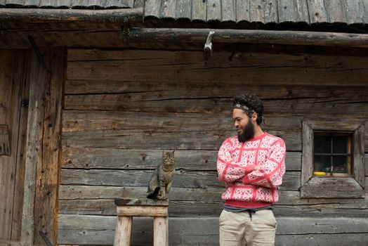 portrait of young hipster man with beard and cat in front of wooden house