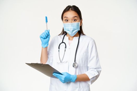 Excited female doctor, asian physician holding clipboard and raising pen up, found solution or idea, standing in medical face mask over white background.