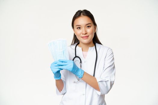 Smiling asian physician, family doctor showing sterile face masks, preventing catching covid-19, standing in uniform over white background.