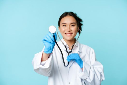 Healthcare and patient checkup concept. Smiling asian woman doctor, nurse examining with stethoscope, standing in medical uniform over blue background.