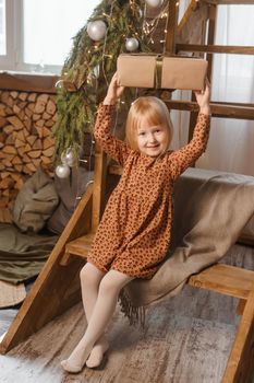 A little blonde girl is sitting on a wooden staircase in a Scandinavian interior decorated in a New Year's style. A child holds a Christmas gift in a craft package.