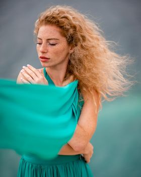 Outdoor portrait of a young beautiful natural redhead girl with freckles, long curly hair, in an emerald dress, posing against the background of the sea