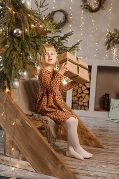 A little blonde girl is sitting on a wooden staircase in a Scandinavian interior decorated in a New Year's style. A child holds a Christmas gift in a craft package.