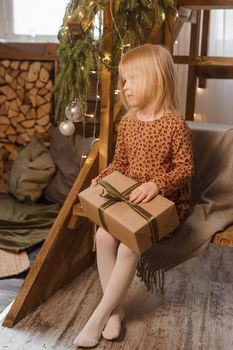A little blonde girl is sitting on a wooden staircase in a Scandinavian interior decorated in a New Year's style. A child holds a Christmas gift in a craft package.