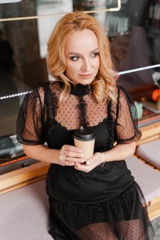 Young charming blonde with a cute smile and makeup while relaxing in a cafe. She is holding a cup of coffee in her hands. She is dressed in a black dress with transparent sleeves