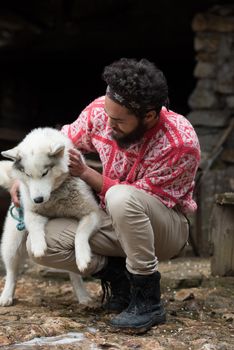 handsome young hipster man standing together with white husky dog in front of old vintage retro wooden house