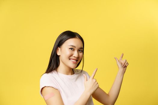 Covid-19 vaccination concept. Cute vaccinated asian woman demonstrating banner, pointing fingers at empty space for promotional text, yellow background.