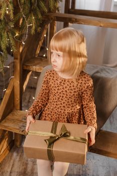 A little blonde girl is sitting on a wooden staircase in a Scandinavian interior decorated in a New Year's style. A child holds a Christmas gift in a craft package.