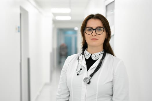 Confident smiling young woman doctor standing in medical institution. Proud professional doctor therapist woman in glasses looking at camera in hospital corridor. Portrait of general practitioner.