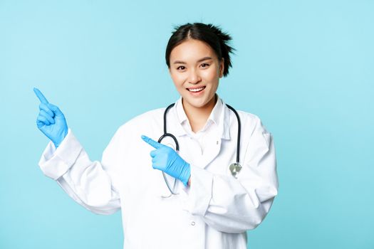 Smiling happy female doctor or nurse, pointing fingers left, wearing medical uniform and gloves, showing hospital advertisement, blue background.