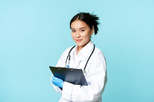 Healthcare and clinic concept. Smiling korean doctor, woman physician in medical uniform, holding clipboard, standing over blue background.